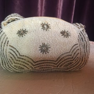 Puede incluir: Un bolso de mano blanco de cuentas con detalles plateados. El bolso está decorado con un patrón repetido de pequeñas estrellas.