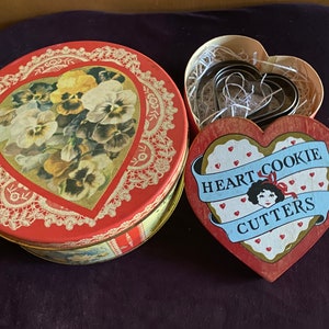 May include: A red heart-shaped tin with a floral design and lace trim. The tin contains a set of heart-shaped cookie cutters. The lid of the tin is open, revealing the cookie cutters and a red heart-shaped box with the words "Heart Cookie Cutters" and a girl's face.