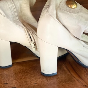May include: Cream-colored leather boots with a block heel and a zipper closure. The boots have a gold-colored decorative accent on the side. The boots are on a wooden surface.