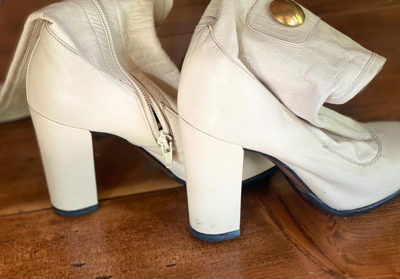 May include: Cream-colored leather boots with a block heel and a zipper closure. The boots have a gold-colored decorative accent on the side. The boots are on a wooden surface.