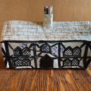 Puede incluir: Una pequeña casa de cerámica con un esmalte blanco y detalles pintados en negro. La casa tiene una chimenea y un techo de paja. La fachada de la casa tiene una puerta negra pintada y ventanas con celosías negras. Hay pequeñas flores moradas pintadas en las enredaderas.