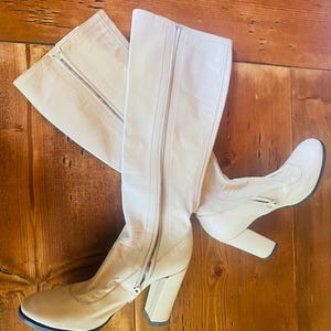 May include: A pair of cream-colored knee-high boots with a zipper closure. The boots have a block heel and a rounded toe. The boots are made of a smooth material and are set against a wooden background.