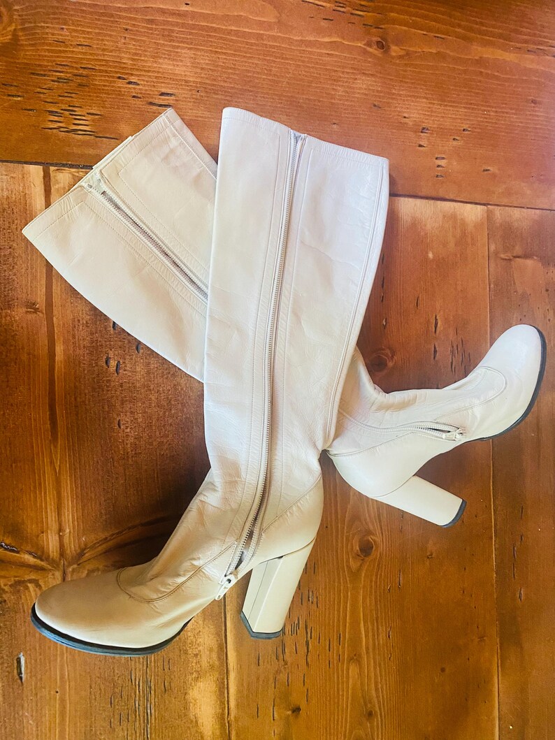May include: A pair of cream-colored knee-high boots with a zipper closure. The boots have a block heel and a rounded toe. The boots are made of a smooth material and are set against a wooden background.