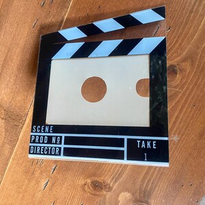 May include: A black and white film clapperboard with the words "SCENE", "PROD NO", "DIRECTOR", and "TAKE 1" printed on it. The clapperboard has a rectangular opening with two circular cutouts. The top has black and white stripes.