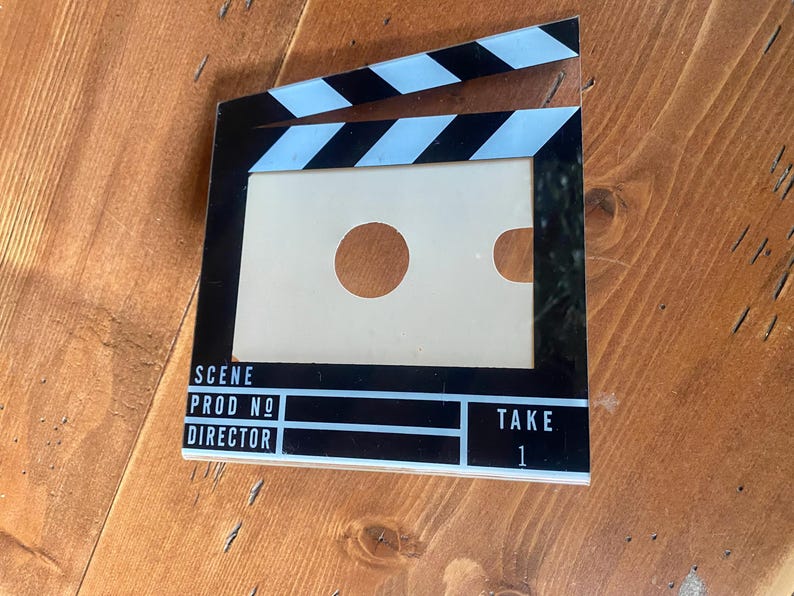 May include: A black and white film clapperboard with the words "SCENE", "PROD NO", "DIRECTOR", and "TAKE 1" printed on it. The clapperboard has a rectangular opening with two circular cutouts. The top has black and white stripes.