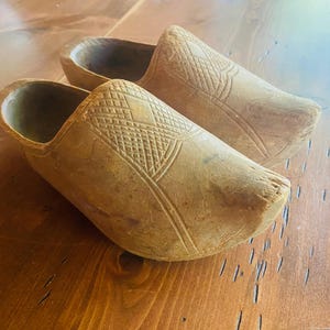 Puede incluir: Un par de zuecos de madera de color marrón claro, con patrones geométricos tallados en la parte superior. Los zuecos se exhiben sobre una superficie de madera, destacando su diseño tradicional y artesanía. Estos zuecos son un artículo de calzado clásico.