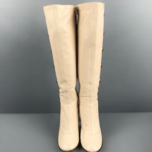 May include: A pair of knee-high cream-colored boots. The boots have a rounded toe and a zipper closure on the inner side. Gold-colored buttons adorn the outer side of the boots. The boots are made of a smooth leather-like material.