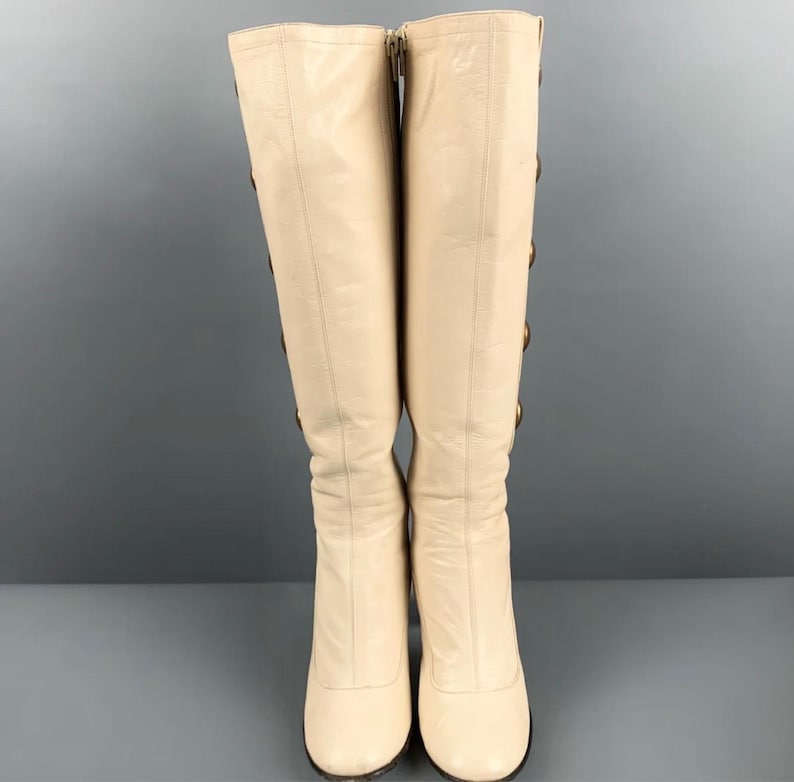 May include: A pair of knee-high cream-colored boots. The boots have a rounded toe and a zipper closure on the inner side. Gold-colored buttons adorn the outer side of the boots. The boots are made of a smooth leather-like material.