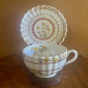 Könnte beinhalten: Ein weißes Tassen- und Untertassen-Set mit einem floralen Design in Gelb, Blau und Rot. Die Tasse hat einen gewellten Rand und steht auf einem Holzsockel. Die Untertasse befindet sich hinter der Tasse.