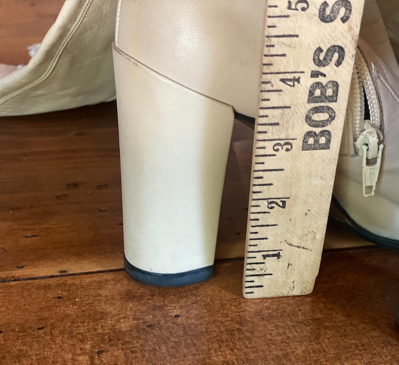May include: Close-up of a cream-colored boot with a chunky heel. A wooden ruler, marked with inches, is placed next to the heel. The heel measures approximately 3 inches in height.