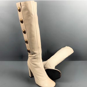 May include: Cream-colored knee-high boots with a block heel. The boots feature a row of large, gold-colored buttons down the side. The boots are made of a smooth material and have a square toe.