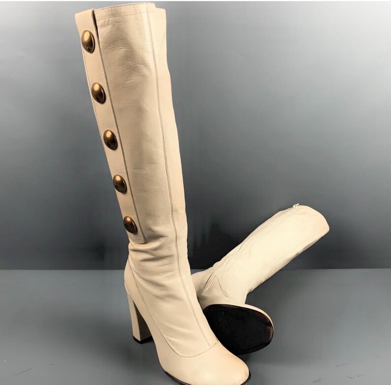 May include: Cream-colored knee-high boots with a block heel. The boots feature a row of large, gold-colored buttons down the side. The boots are made of a smooth material and have a square toe.