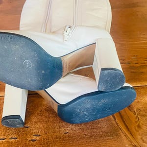 May include: A pair of white leather knee-high boots with block heels. The boots have a zipper closure and a blue sole. The heels are approximately 4 inches high.