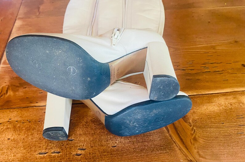 May include: A pair of white leather knee-high boots with block heels. The boots have a zipper closure and a blue sole. The heels are approximately 4 inches high.