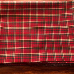 May include: Folded red plaid fabric with a pattern of black, green, and cream-colored lines. The fabric is folded neatly, showing the pattern clearly. The plaid design is a classic pattern, often used for home decor and textiles.