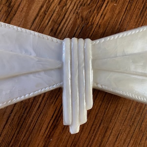 以下が含まれることがあります： ロープ状のテクスチャのある縁取りが施された、白の真珠光沢のあるプラスチック製の蝶ネクタイのブローチ。蝶ネクタイは、複数のプラスチック層で構成されており、重ねられた効果を生み出しています。