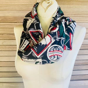 May include: A black, white, red, and green infinity scarf with a vintage travel-themed print. The scarf features various travel-related graphics, including the word "HOTEL" and the name "DEAUVILLE". The scarf is displayed on a white mannequin.