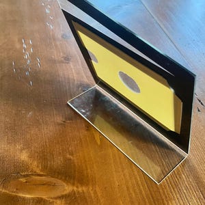 May include: A clear acrylic photo frame with a black border, angled on a wooden surface. The frame holds a yellow insert with two oval cutouts. The frame is designed to stand upright.