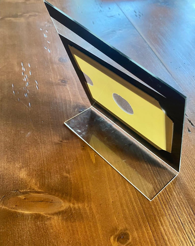 May include: A clear acrylic photo frame with a black border, angled on a wooden surface. The frame holds a yellow insert with two oval cutouts. The frame is designed to stand upright.