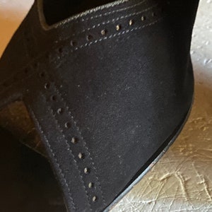 Op de afbeelding: Close-up van een zwarte su&egrave;de schoen met een decoratief stikselpatroon. De schoen heeft een spitse neus en een hoge hak.