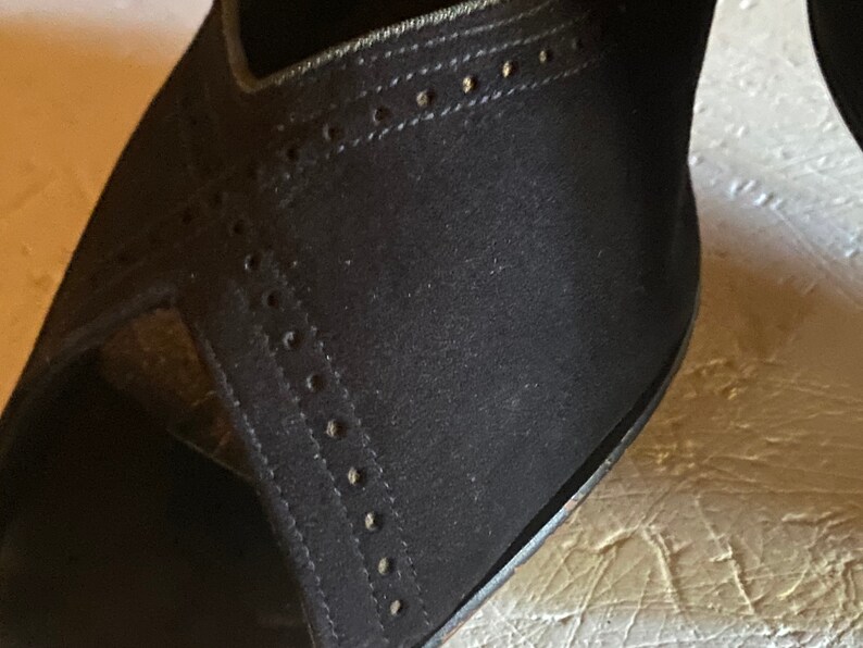 Op de afbeelding: Close-up van een zwarte su&egrave;de schoen met een decoratief stikselpatroon. De schoen heeft een spitse neus en een hoge hak.