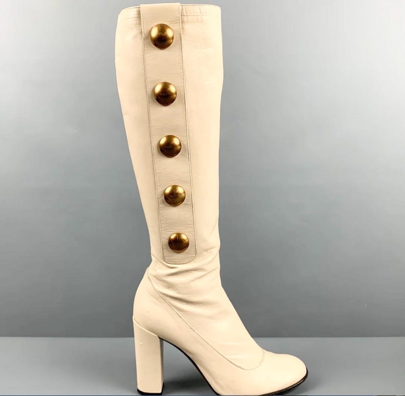 May include: Ivory-colored knee-high boot with a block heel. The boot features a series of large, gold-toned buttons down the side. The leather boot has a rounded toe and a sturdy heel.