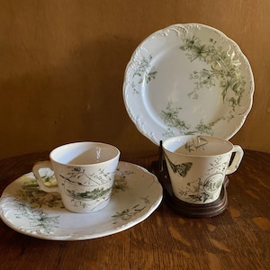 Może przedstawiać: Zestaw vintage filiżanek do herbaty i spodków z zielonym wzorem kwiatowym. Filiżanki i spodki są białe z delikatnym zielonym wzorem kwiatowym. Filiżanki mają zaokrąglony kształt i mały uchwyt. Spodki są nieco większe od filiżanek i mają falisty brzeg.