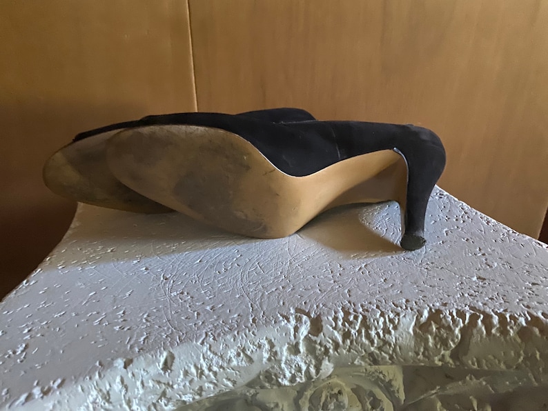 Op de afbeelding: Een paar zwarte su&egrave;de hakken met een spitse neus. De schoenen staan op een wit oppervlak.