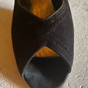 Op de afbeelding: Zwarte su&egrave;de peeptoe hakken met een gouden rand en een decoratief patroon van kleine gaatjes.