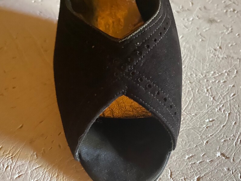 Op de afbeelding: Zwarte su&egrave;de peeptoe hakken met een gouden rand en een decoratief patroon van kleine gaatjes.