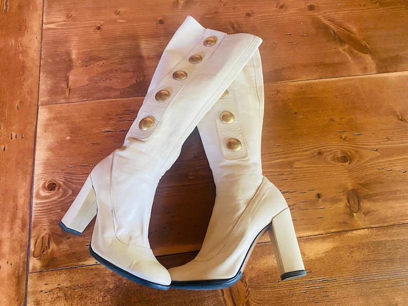 May include: A pair of cream-colored knee-high boots with chunky heels. The boots feature gold-colored button details along the side and a black sole. The boots are made of a smooth material and are set against a wooden background.