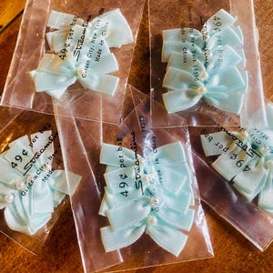May include: A collection of light blue satin bows, each with a pearl centre, packaged in clear plastic bags. The bags are labelled with the brand "Streamline" and "Made in China". These bows are suitable for craft projects.
