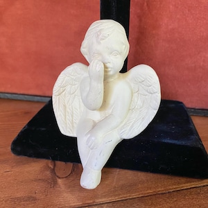 Può includere: Piccola statuina di cherubino bianco sporco, con ali dettagliate, seduto con un dito che tocca il suo viso. Il cherubino è su una superficie di velluto nero, con una superficie di legno sullo sfondo.