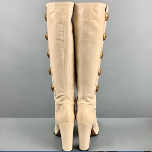 May include: A pair of cream-colored knee-high boots. Each boot features a series of large, gold-toned buttons running down the side. The boots have a block heel and a smooth leather texture, offering a vintage aesthetic.