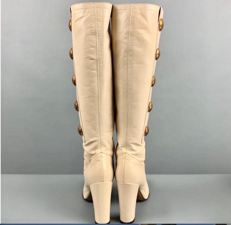 May include: A pair of cream-colored knee-high boots. Each boot features a series of large, gold-toned buttons running down the side. The boots have a block heel and a smooth leather texture, offering a vintage aesthetic.