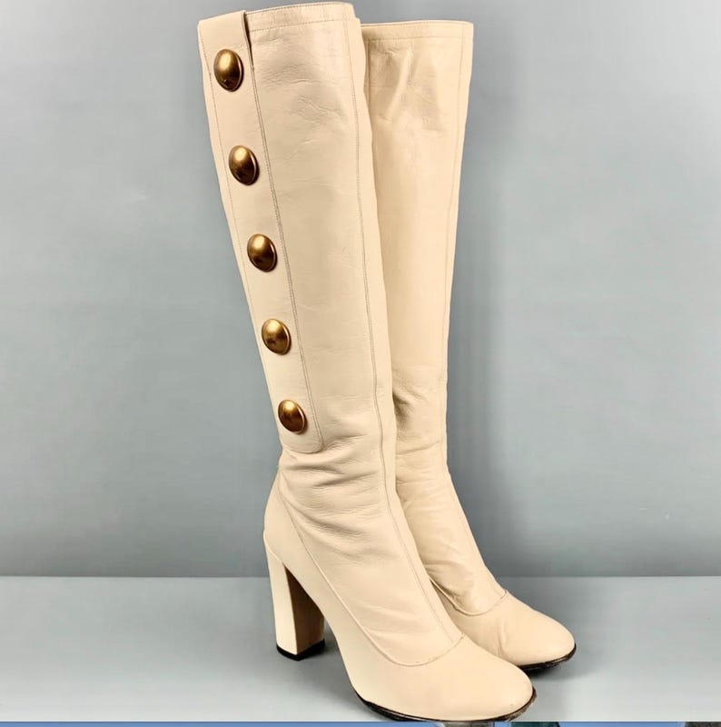 May include: A pair of cream-colored knee-high boots with large gold-tone buttons down the side. The boots have a block heel and a rounded toe. The background is a solid light blue.