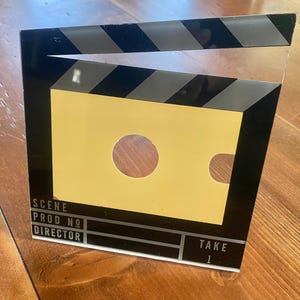 May include: A film clapperboard with a yellow center and black and white stripes at the top. The clapperboard has the words "SCENE", "PROD NO", "DIRECTOR", and "TAKE 1" printed on it. The clapperboard is a prop used in filmmaking.