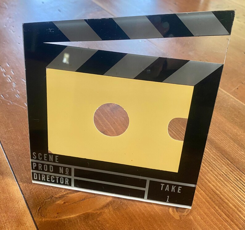 May include: A film clapperboard with a yellow center and black and white stripes at the top. The clapperboard has the words "SCENE", "PROD NO", "DIRECTOR", and "TAKE 1" printed on it. The clapperboard is a prop used in filmmaking.