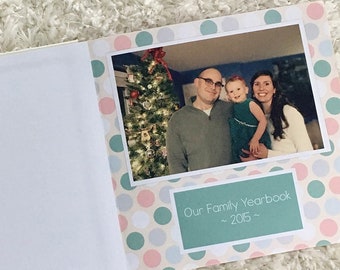 Puede incluir: Una portada de álbum de fotos familiar con un patrón de lunares verde, blanco y rosa. La portada presenta una foto de una familia de tres personas de pie frente a un árbol de Navidad. El texto "Our Family Yearbook ~ 2015 ~" está impreso en la portada.