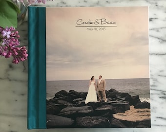 May include: A teal blue wedding album with a photo of a bride and groom standing on rocks by the ocean. The album cover has the text "Coralee & Brian" and "May 18, 2013" printed on it.
