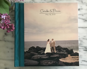 May include: Teal blue wedding album with a photo of a couple on a rocky beach. The album cover has the text "Coralee & Brian" and "May 18, 2013".