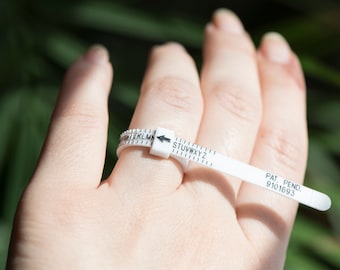 Pode incluir: Um medidor de tamanho de anel de plástico branco com uma seta preta e letras de A a Z. O medidor está no dedo de uma pessoa. O texto "PAT. PEND. 9101693" está impresso no medidor.