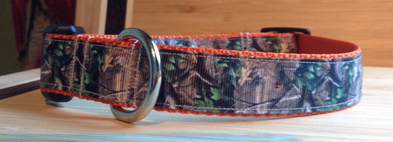 May include: A camouflage dog collar with an orange trim and a silver D-ring.