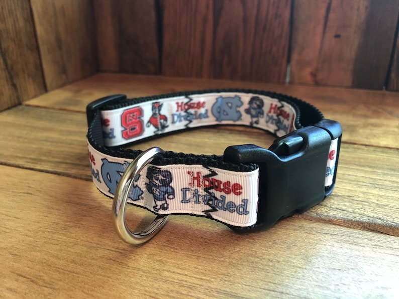 K&ouml;nnte beinhalten: Ein wei&szlig;es Hundehalsband mit einer schwarzen Schnalle und einem silbernen Ring. Das Halsband hat ein sich wiederholendes Muster mit den Worten "House Divided" und Bildern der Maskottchen der University of North Carolina und der North Carolina State University.