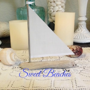 Puede incluir: Un peque&ntilde;o velero blanco hecho de madera a la deriva con una vela blanca. El velero est&aacute; sentado sobre un mantel de crochet blanco con una concha a su lado. El texto "Sweet Beaches" est&aacute; escrito en cursiva azul en el mantel.