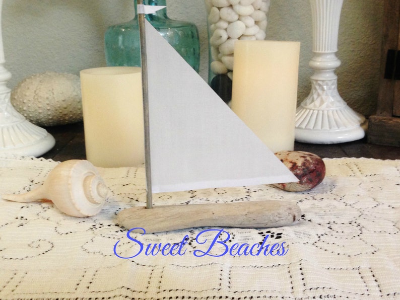 Puede incluir: Un peque&ntilde;o velero blanco hecho de madera a la deriva con una vela blanca. El velero est&aacute; sentado sobre un mantel de crochet blanco con una concha a su lado. El texto "Sweet Beaches" est&aacute; escrito en cursiva azul en el mantel.