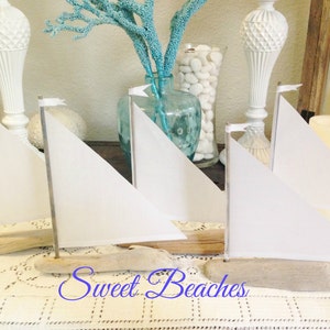 Puede incluir: Tres veleros blancos hechos de madera flotante y tela, con banderas blancas en la parte superior, sentados sobre un mantel de encaje blanco. Los veleros est&aacute;n dispuestos en fila, con el velero m&aacute;s grande en la parte posterior y el velero m&aacute;s peque&ntilde;o en la parte delantera. Los veleros est&aacute;n decorados con velas de tela blanca y banderas blancas. Los veleros est&aacute;n sentados sobre un mantel de encaje blanco. Los veleros son una decoraci&oacute;n de tem&aacute;tica n&aacute;utica.