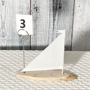 May include: A small wooden sailboat with a white sail and a number "3" on a card attached to the front. The sailboat is on a white textured surface.
