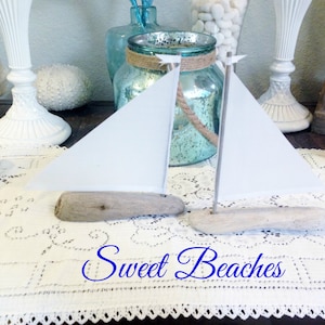 Puede incluir: Dos veleros blancos hechos de madera a la deriva y papel, con un mantel de encaje blanco y el texto "Sweet Beaches" en cursiva azul.