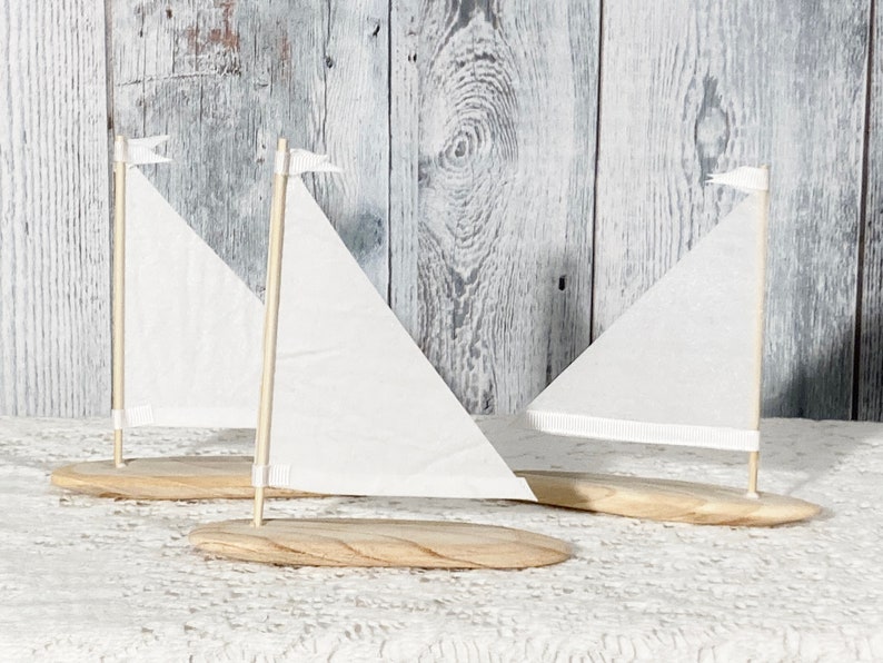 Puede incluir: Tres veleros de madera en miniatura con velas blancas y banderas blancas en la parte superior. Los veleros est&aacute;n sobre un mantel de encaje blanco.